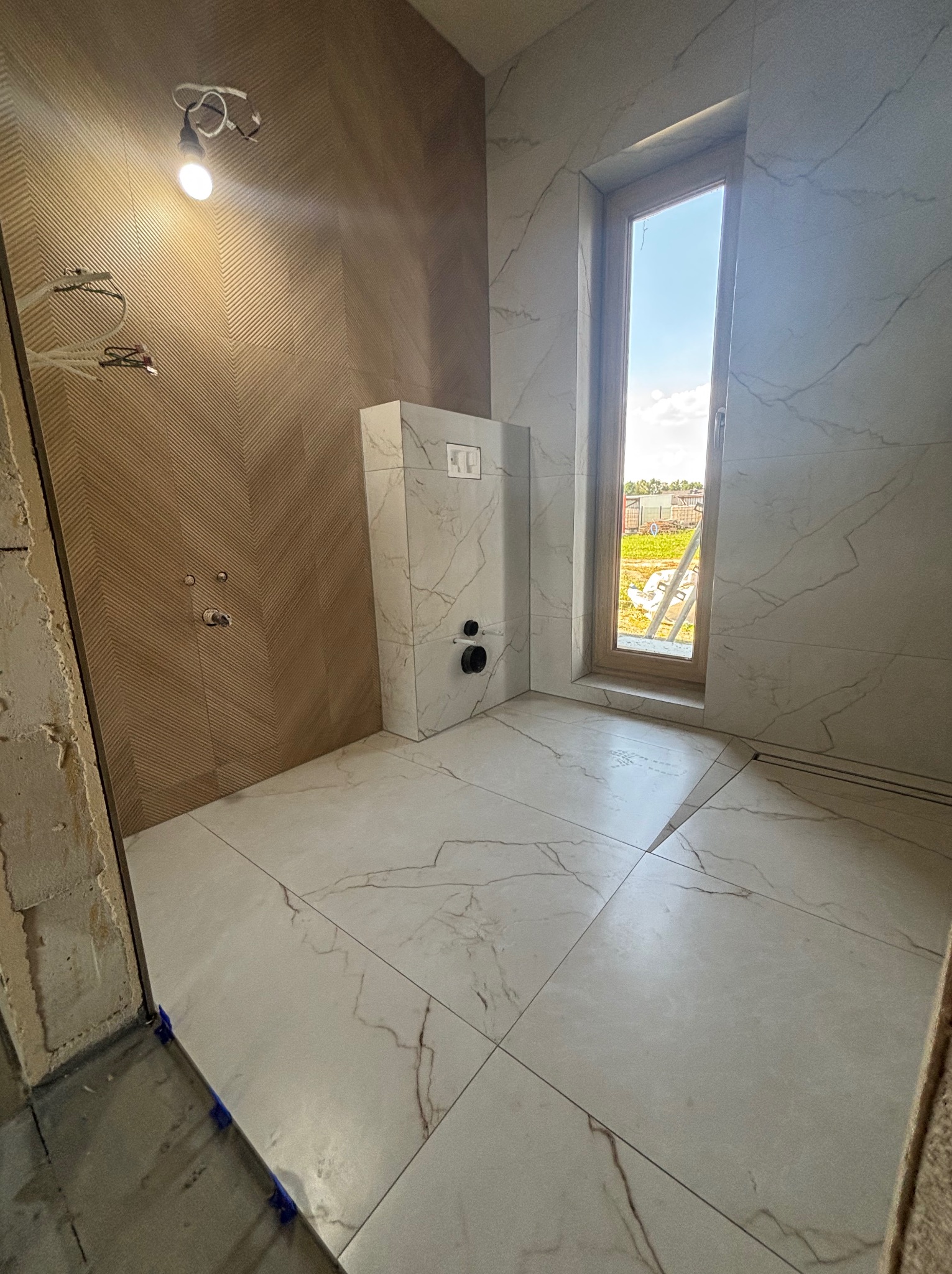 Łazienka w trakcie wykańczania: ułożone płytki podłogowe imitujące marmur, ściana z brązowymi płytkami w jodełkę, okno z widokiem na zewnątrz, częściowo wykończona ściana.
