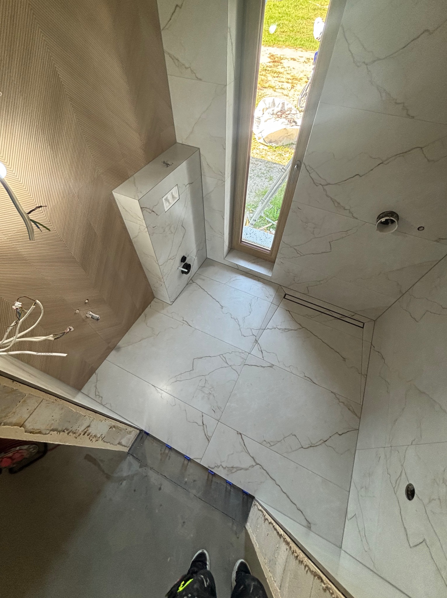 Wykończona łazienka z białymi płytkami imitującymi marmur, widok z góry na podłogę z odpływem liniowym i fragment ściany z drewnopodobną fakturą.