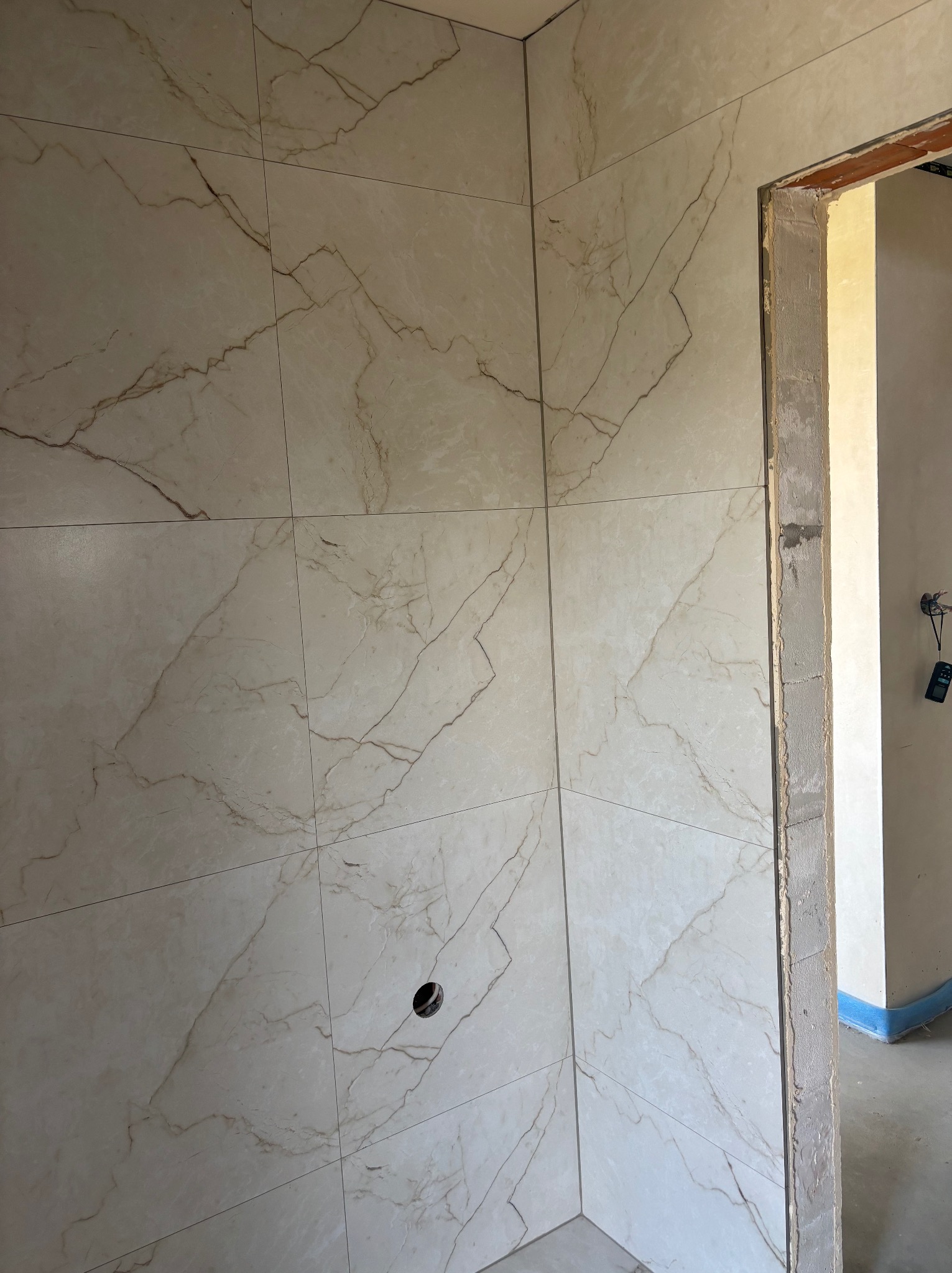 Wykończona ściana z płytek imitujących marmur w łazience, widoczny otwór na instalację i surowa ościeżnica drzwiowa. Jasne wnętrze w trakcie prac wykończeniowych.