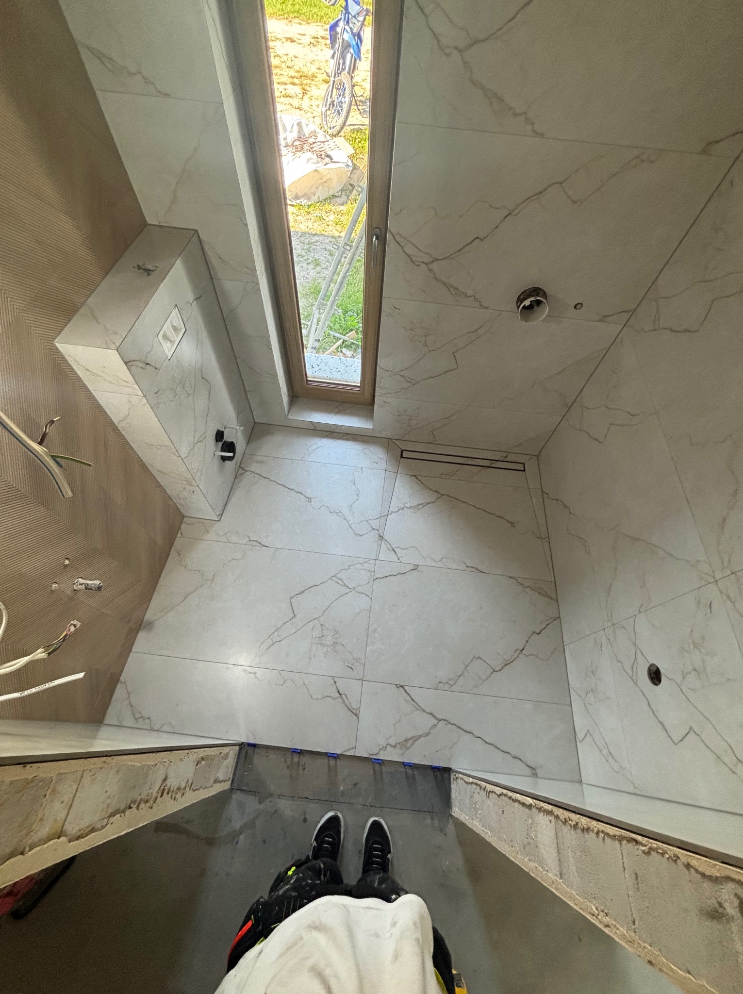 Wykończona łazienka z marmurowymi płytkami, widok z góry na podłogę i fragment osoby. Uwagę przykuwa okno dachowe i minimalistyczny odpływ liniowy.