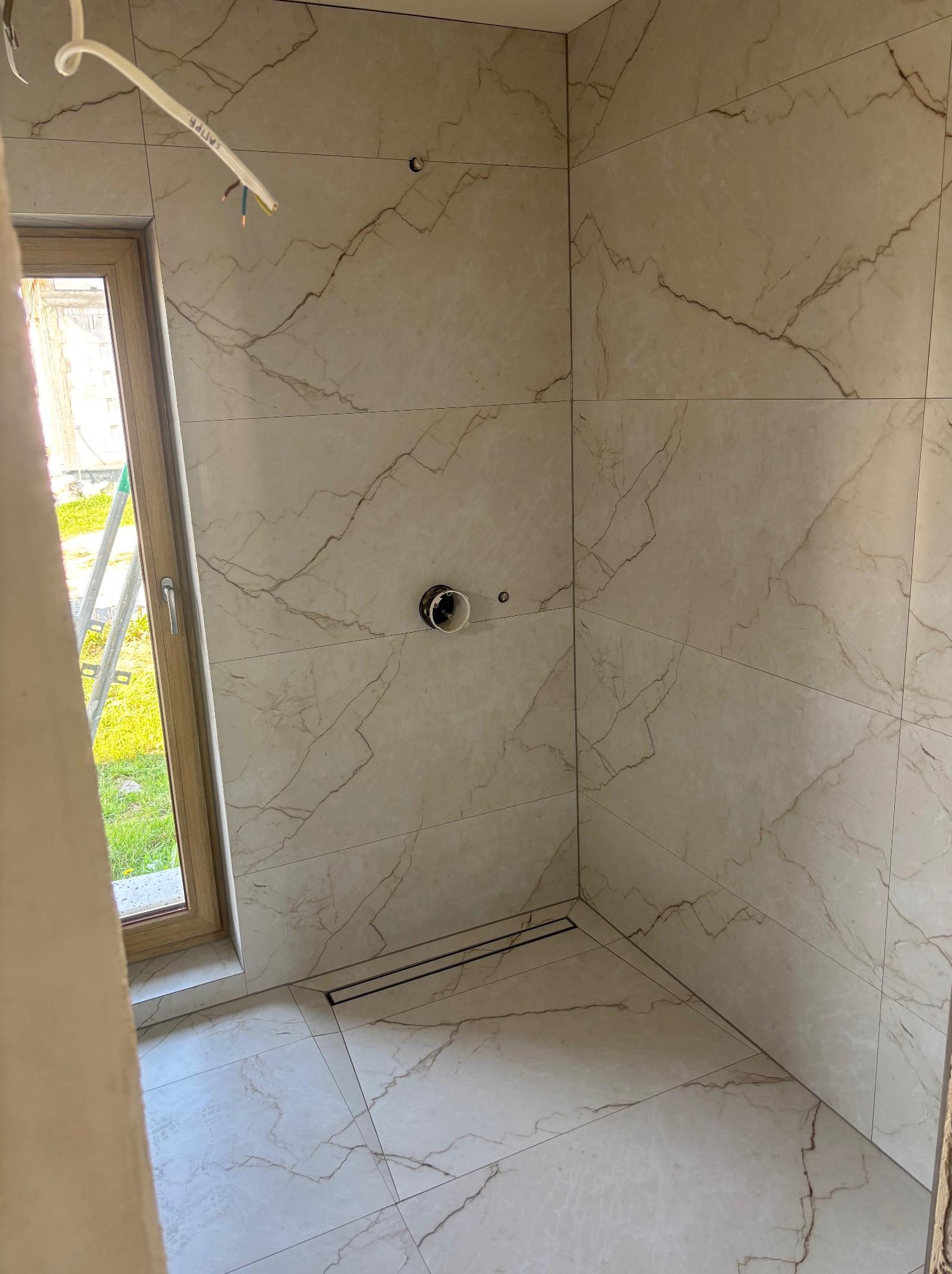 Wykończona kabina prysznicowa z beżowymi płytkami imitującymi marmur, z odpływem liniowym i oknem z widokiem na zieleń. Widoczne przewody elektryczne.