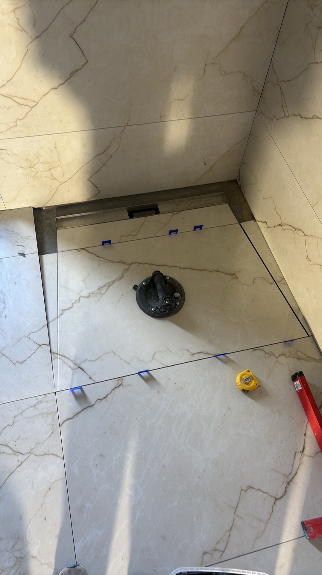 Układanie płytek imitujących marmur w łazience. Widoczna przyssawka do płytek, poziomica i miarka. Użyto niebieskich krzyżyków dystansowych.