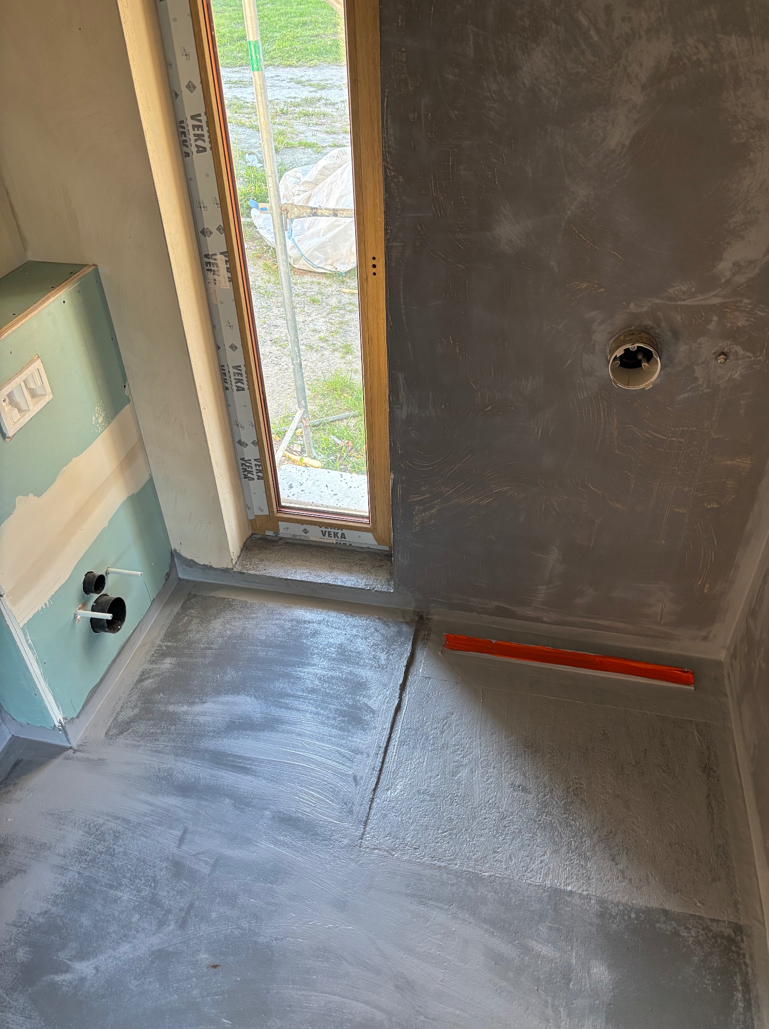 Łazienka w trakcie remontu: hydroizolacja podłogi i ścian, widoczne rury odpływowe, okno z profilem Veka, poziomica na podłodze. Szare wykończenie.