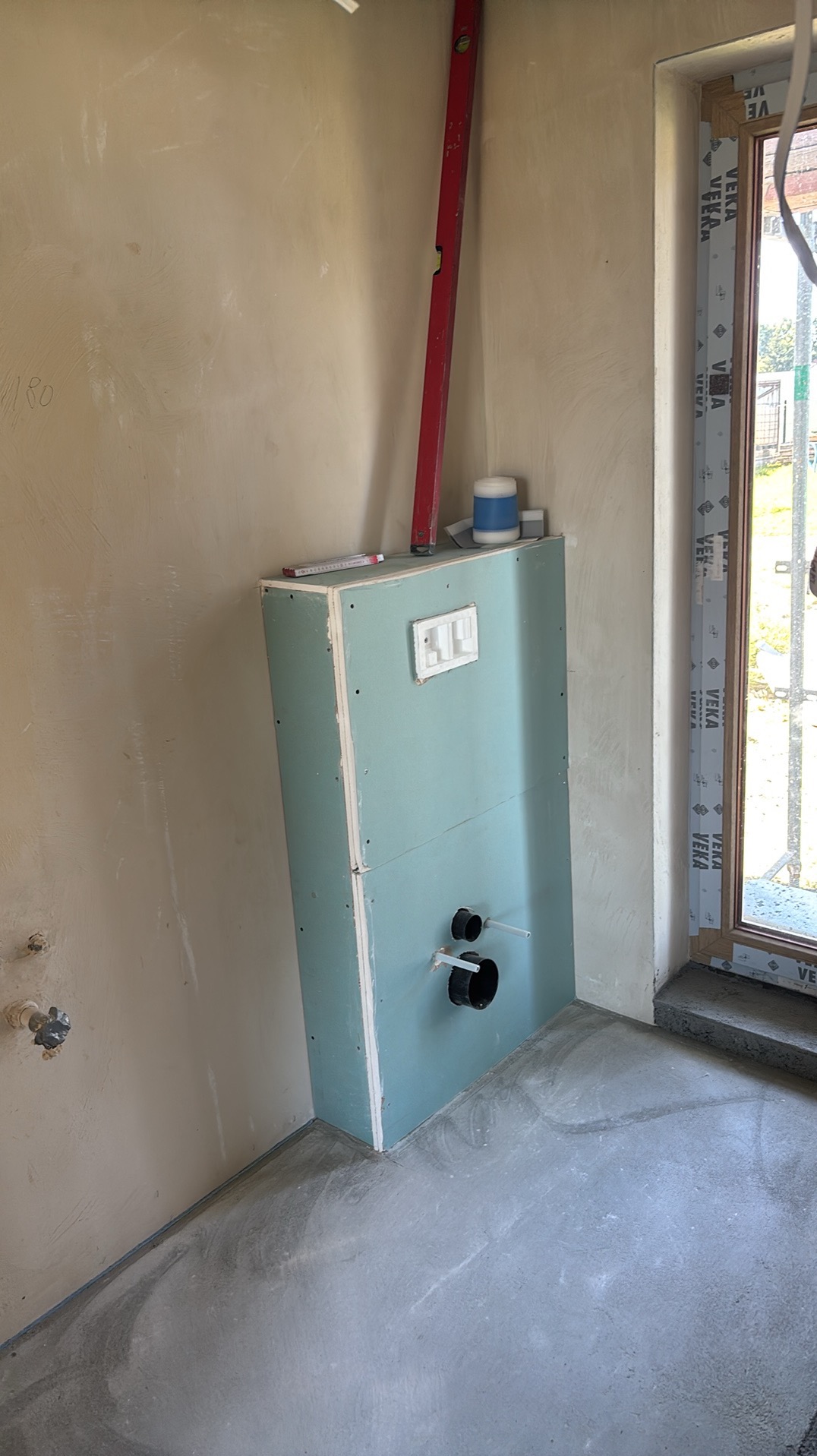 Stelaż podtynkowy zabudowany płytą gipsowo-kartonową w surowej łazience, widoczne przyłącza kanalizacyjne i wodne, poziomica oparta o ścianę, okno z PCV w tle.