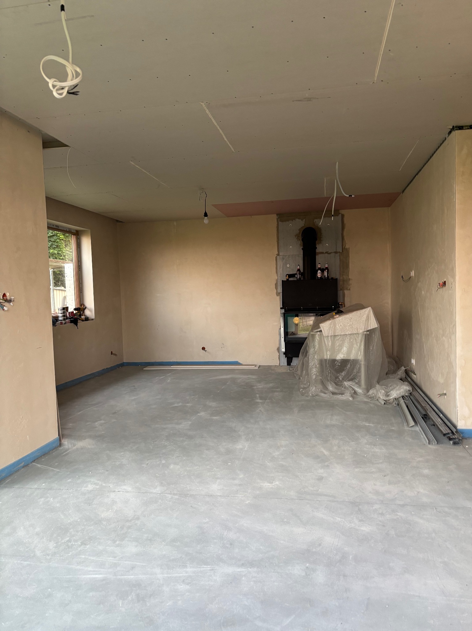 Wnętrze w trakcie prac wykończeniowych: surowe ściany, wylewka podłogowa, kominek zabezpieczony folią. Widoczne przewody elektryczne i materiały budowlane.