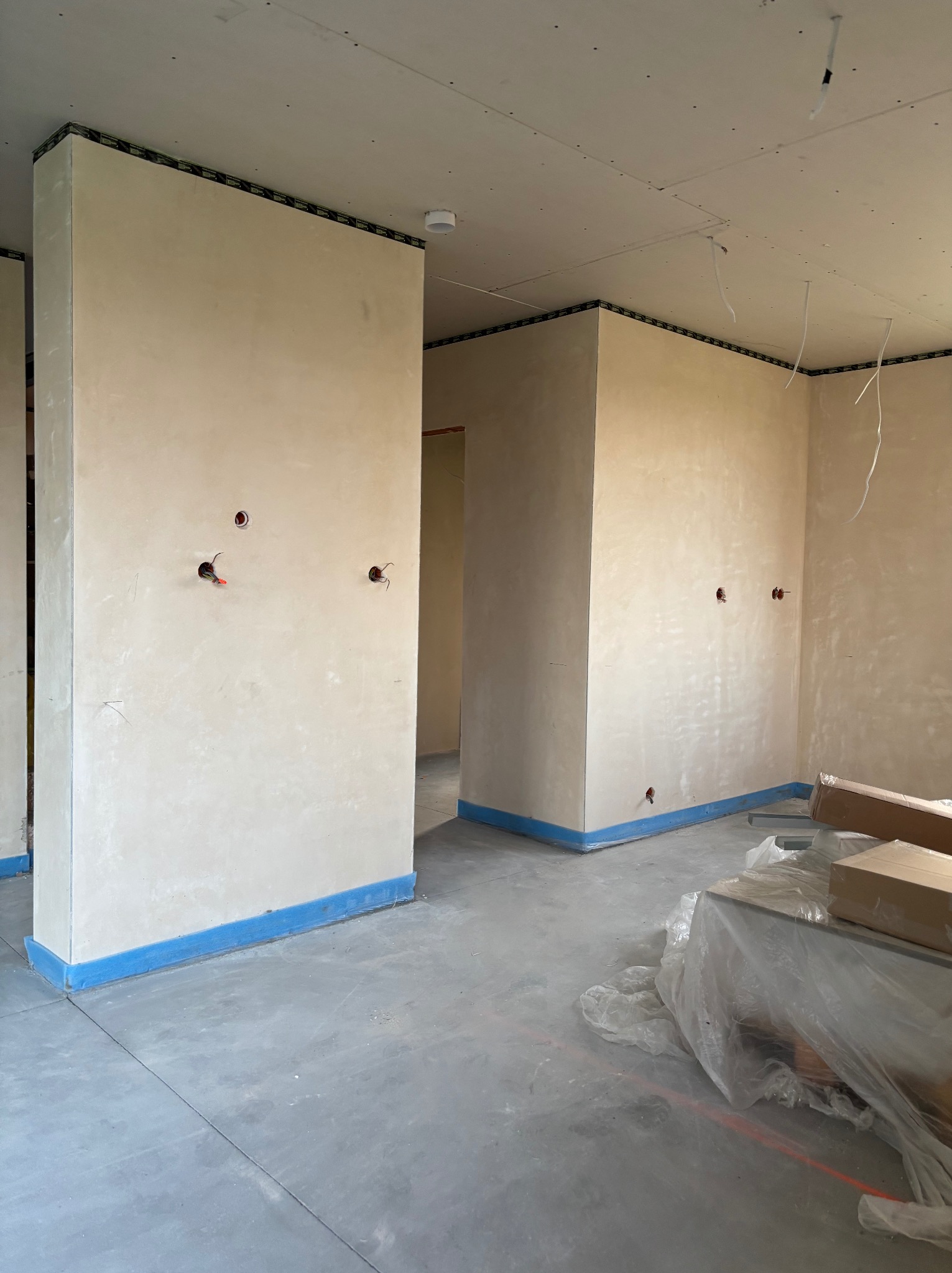 Wnętrze w trakcie prac wykończeniowych: ściany po szpachlowaniu, zabezpieczone taśmą malarską, widoczne puszki elektryczne, sufit z płyt karton-gips.
