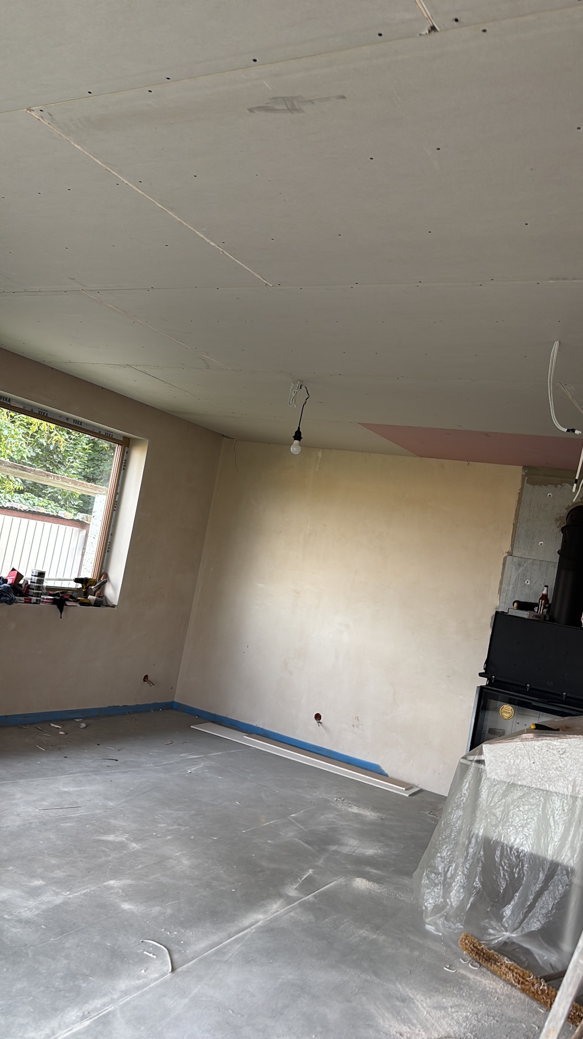 Wnętrze w trakcie prac wykończeniowych: ściany po szpachlowaniu, sufit z płyt karton-gips, okno z widokiem na zieleń. Widoczne narzędzia i materiały budowlane.