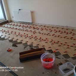 Kuba kępski - Układanie parkietu w jodełkę z użyciem klipsów poziomujących. Widoczne narzędzia i materiały. Częściowo ułożona podłoga w jasnym pomieszczeniu z grzejnikiem.