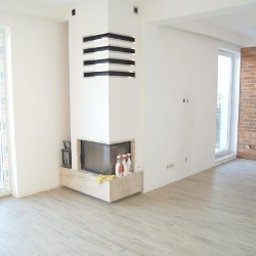 Kuba kępski - Jasne, przestronne wnętrze po remoncie: białe ściany, drewniana podłoga, ceglana ściana akcentująca, nowoczesny kominek narożny z dekoracyjnymi elementami.