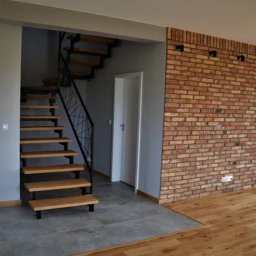 Kuba kępski - Nowoczesne schody drewniane z czarną balustradą, ceglana ściana i szara podłoga w jasnym wnętrzu. Widoczne przejście do innego pomieszczenia.