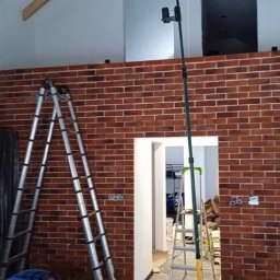 Kuba kępski - Ściana z cegły klinkierowej w trakcie montażu, widoczne drabiny i narzędzia pomiarowe. Częściowo wykończone wnętrze z widocznym otworem drzwiowym.