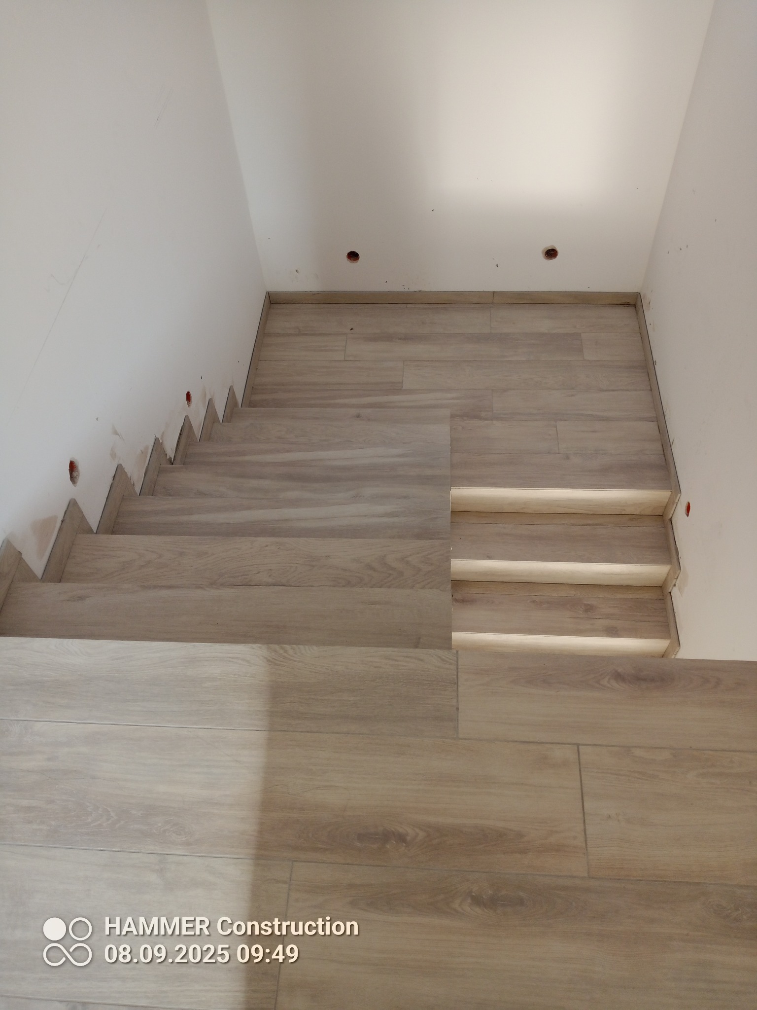 Wykończone schody z drewnopodobnych płytek, widoczne otwory na oświetlenie w ścianie, surowy stan wykończenia ściany przy schodach. Ujęcie z góry.