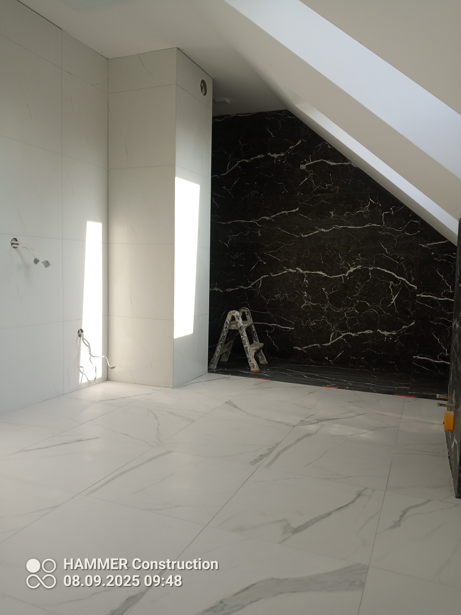 Wykończona łazienka na poddaszu z białymi płytkami imitującymi marmur na ścianach i podłodze, oraz czarną ścianą z wzorem marmuru. Widoczna drabina.