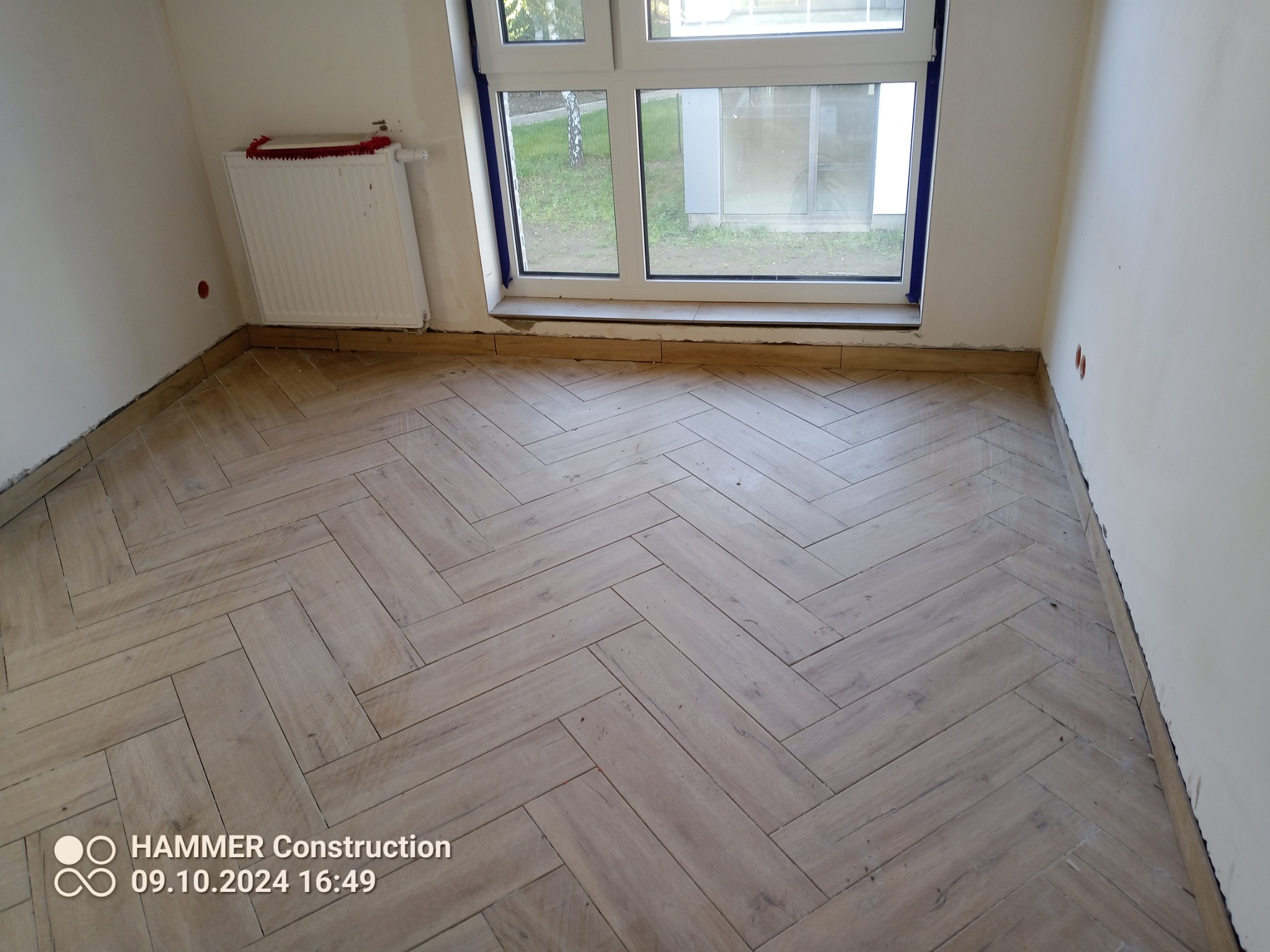 Ukończona podłoga z paneli ułożonych w jodełkę w jasnym pomieszczeniu z oknem. Widoczny grzejnik i fragmenty ścian przed wykończeniem.