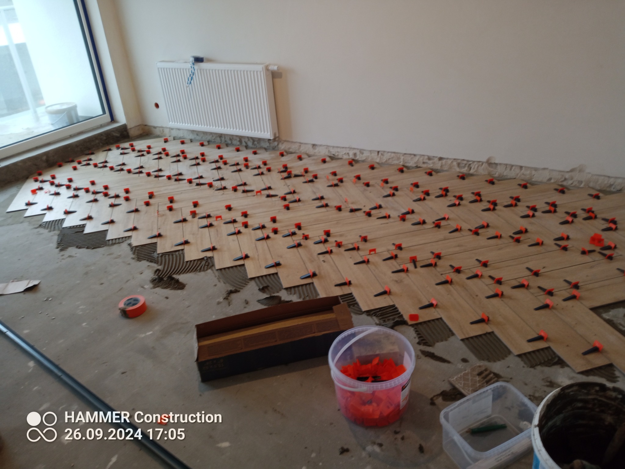 Układanie parkietu w jodełkę z użyciem klipsów poziomujących. Widoczne narzędzia i materiały. Częściowo ułożona podłoga w jasnym pomieszczeniu z grzejnikiem.