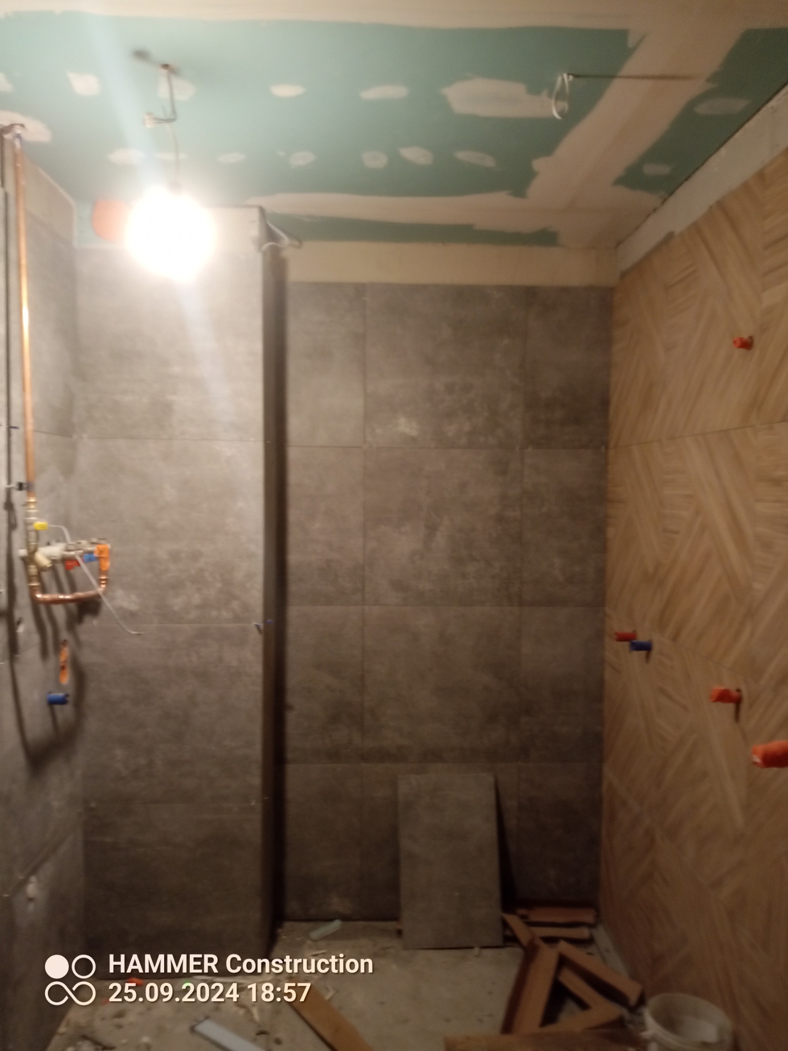 Wnętrze łazienki w trakcie remontu: częściowo ułożone płytki w odcieniach szarości i drewna, widoczne instalacje hydrauliczne i fragment sufitu z płyt gipsowych.