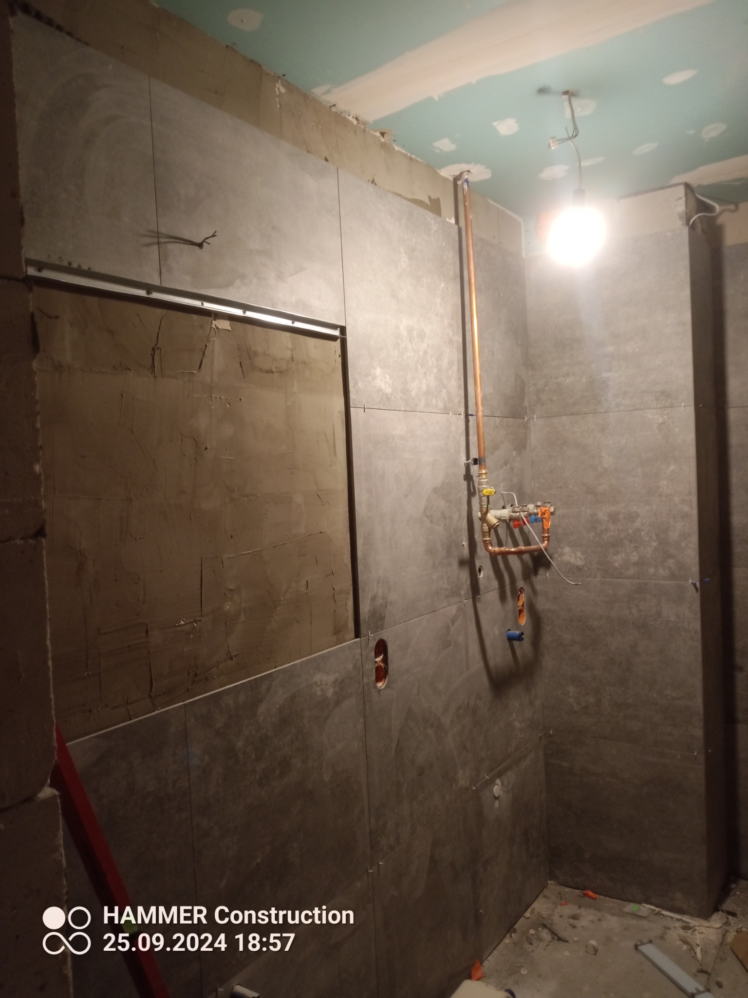 Montaż płytek w łazience z widoczną instalacją hydrauliczną. Szare płytki częściowo ułożone, widoczne krzyżyki dystansowe. Fragment ściany przygotowany pod płytki.