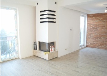 Jasne, przestronne wnętrze po remoncie: białe ściany, drewniana podłoga, ceglana ściana akcentująca, nowoczesny kominek narożny z dekoracyjnymi elementami.