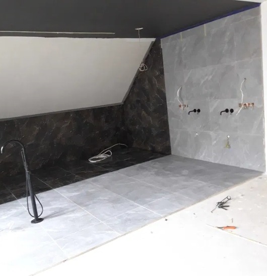 Wykończona łazienka z szarymi i brązowymi płytkami imitującymi marmur, czarną armaturą i narzędziami na podłodze. Ukośny sufit pomalowany na ciemny kolor.