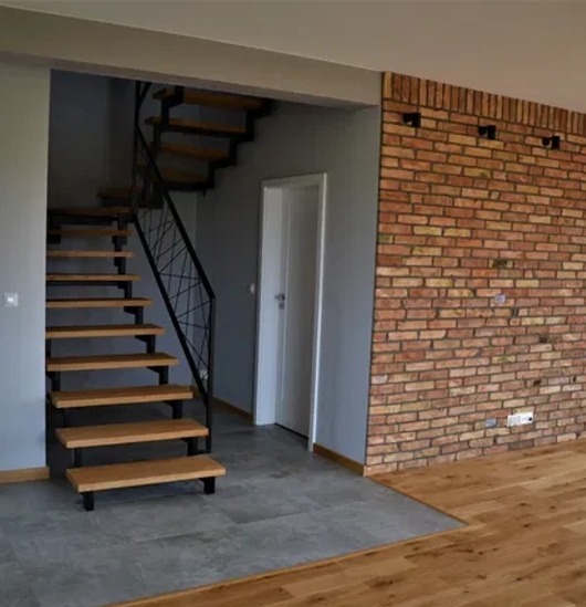 Nowoczesne schody drewniane z czarną balustradą, ceglana ściana i szara podłoga w jasnym wnętrzu. Widoczne przejście do innego pomieszczenia.