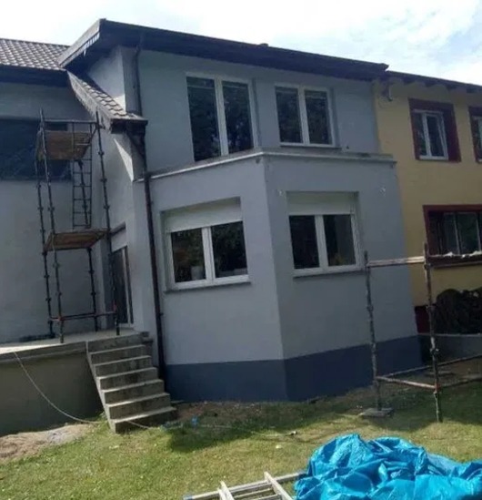 Szary dom po malowaniu elewacji z rusztowaniem, widok z ogrodu. Dolna część elewacji w ciemniejszym odcieniu szarości. Okna z białymi ramami.
