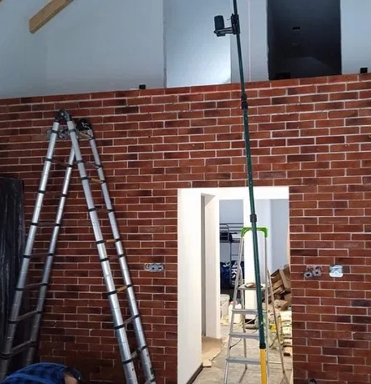 Ściana z cegły klinkierowej w trakcie montażu, widoczne drabiny i narzędzia pomiarowe. Częściowo wykończone wnętrze z widocznym otworem drzwiowym.