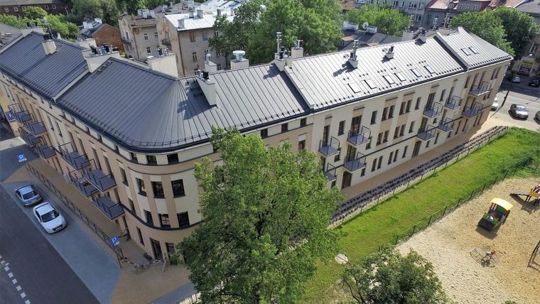 Widok z góry na odnowiony budynek wielorodzinny z szarą blachą na dachu, małymi balkonami i placem zabaw w tle. Dwa zaparkowane samochody.