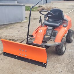 TRZYNASTKA ADRIAN SZCZECHOWSKI - Pomarańczowy traktorek z pługiem śnieżnym Husqvarna na betonowym podłożu. Widoczne detale mocowania pługu i opony z bieżnikiem. Sprzęt gotowy do pracy.