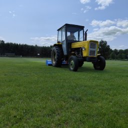 TRZYNASTKA ADRIAN SZCZECHOWSKI - Żółty traktor z kabiną kosi trawę na zielonym boisku sportowym w słoneczny dzień. W tle widoczne drzewa i błękitne niebo z chmurami.