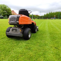 TRZYNASTKA ADRIAN SZCZECHOWSKI - Pomarańczowa kosiarka traktorek Husqvarna na idealnie skoszonym, zielonym trawniku. W tle widoczne drzewa i elementy placu zabaw. Perfekcyjne koszenie.