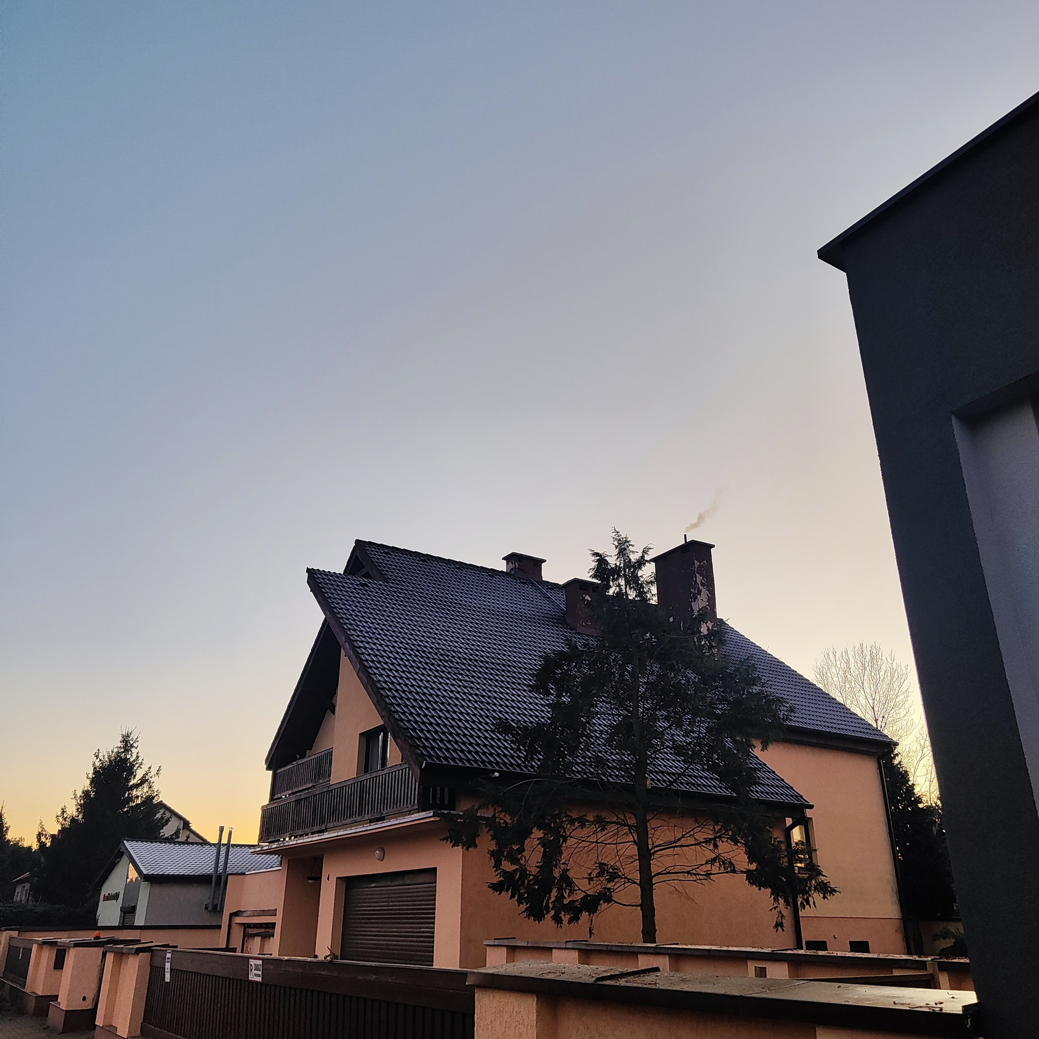 Dwupiętrowy dom z ciemnym dachem i balkonem, widziany z dołu pod kątem. Na dachu komin z dymem. W tle błękitne niebo o zachodzie słońca. Z przodu drzewo.
