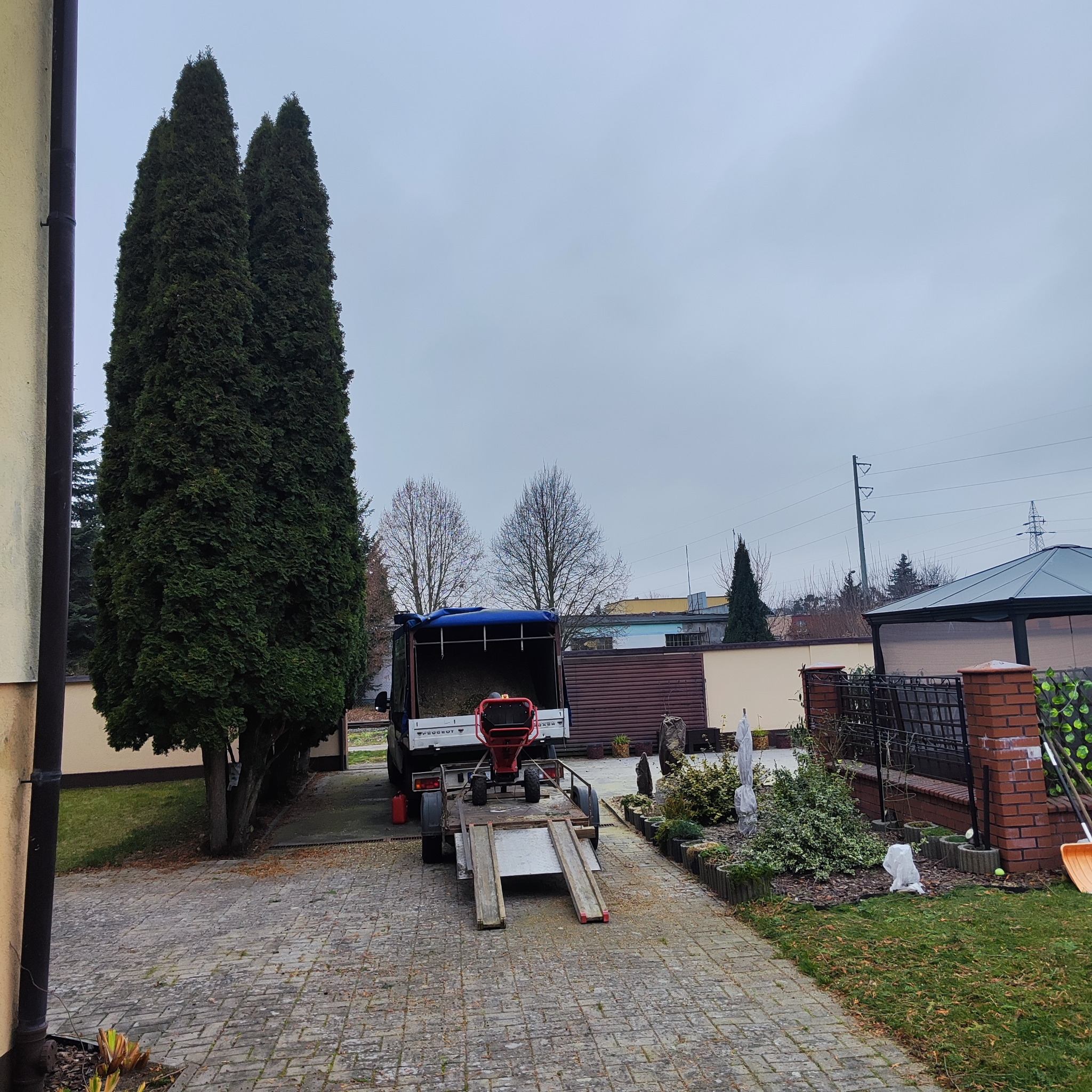 Ciężarówka z przyczepą i rębakiem do gałęzi na posesji z tujami. Sprzęt gotowy do pracy przy drzewach i krzewach. Widok na podwórko z kostki.
