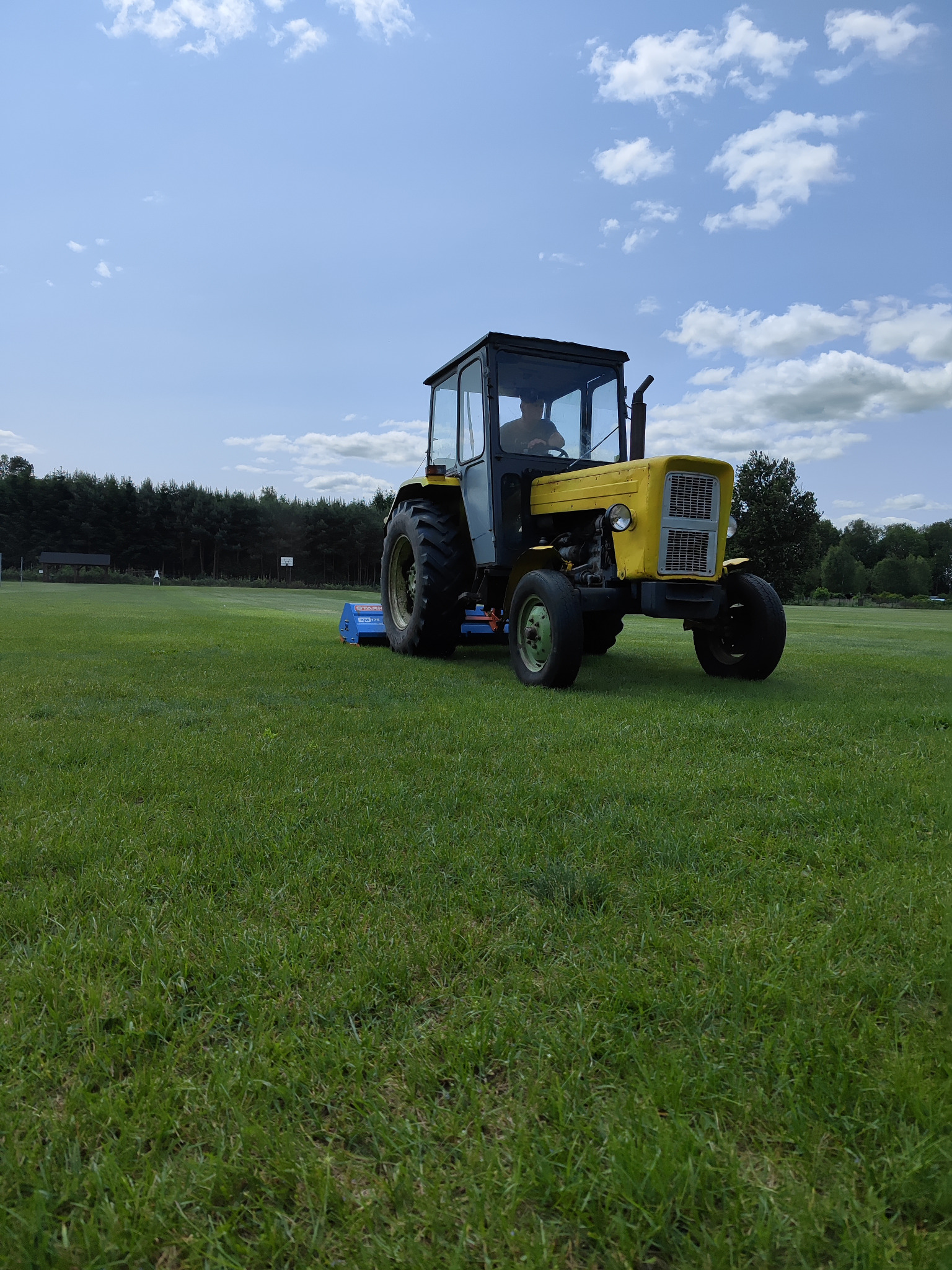 Żółty traktor z kabiną kosi trawę na zielonym boisku sportowym w słoneczny dzień. W tle widoczne drzewa i błękitne niebo z chmurami.