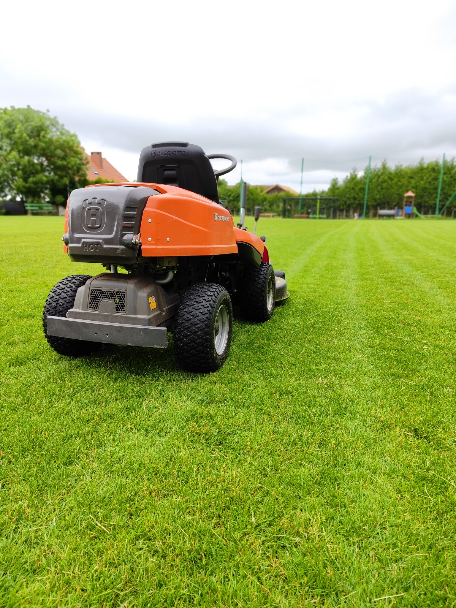 Pomarańczowa kosiarka traktorek Husqvarna na idealnie skoszonym, zielonym trawniku. W tle widoczne drzewa i elementy placu zabaw. Perfekcyjne koszenie.