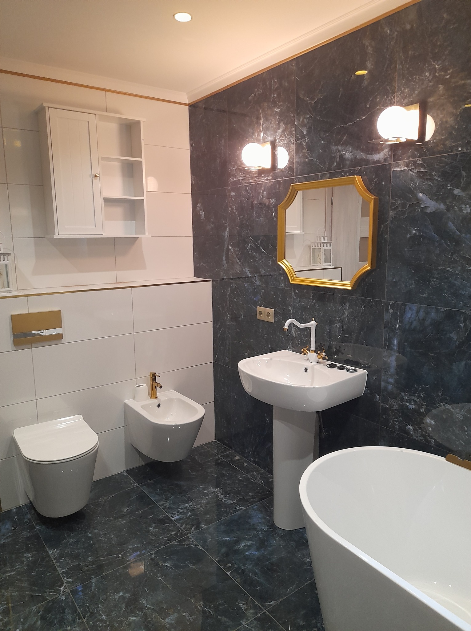 Elegancka łazienka z marmurowymi płytkami, złotymi akcentami i nowoczesną armaturą: umywalka na postumencie, toaleta, bidet i wanna. Lustro w złotej ramie.