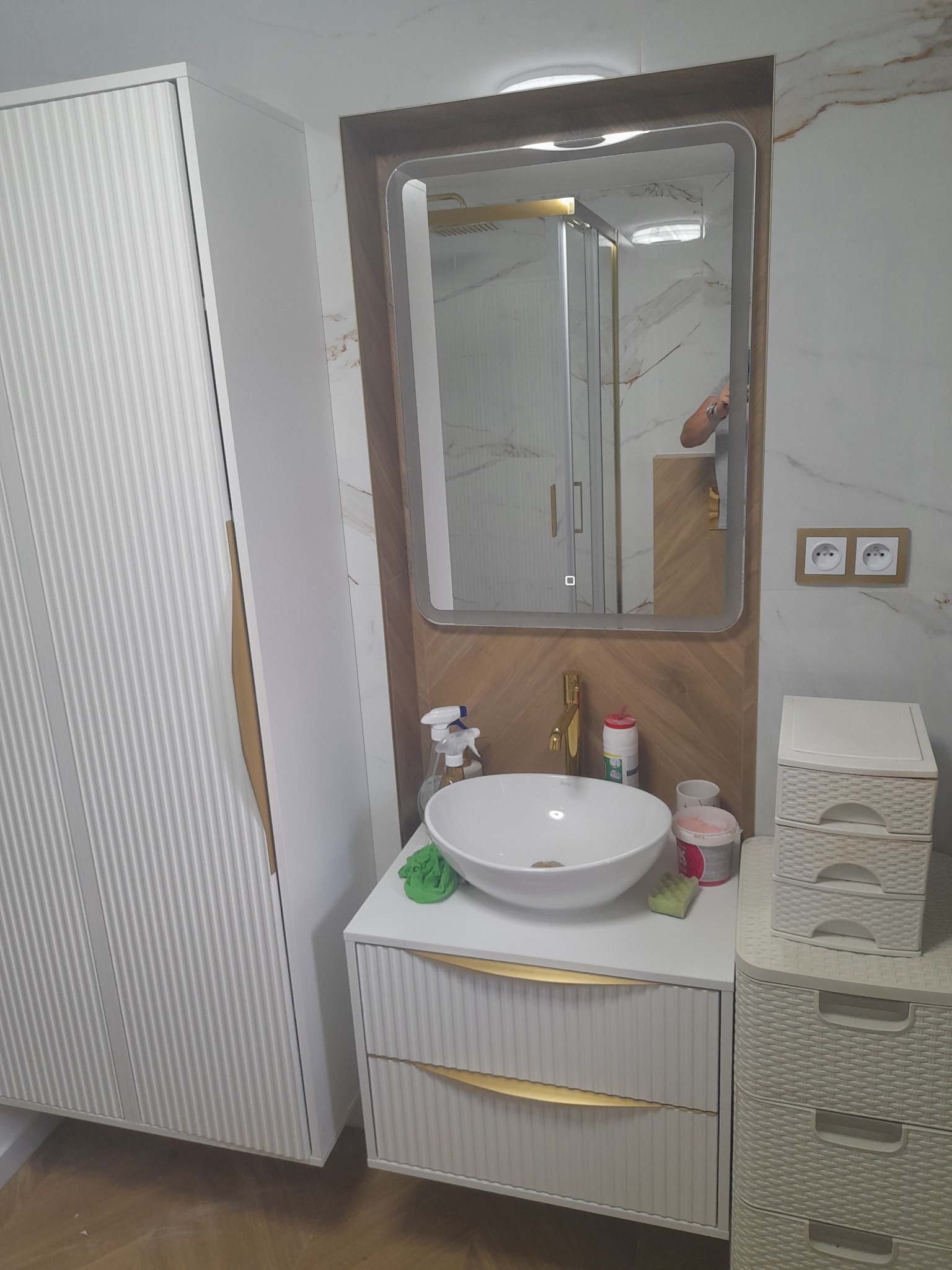 Nowoczesna łazienka z białą szafką pod umywalką, złotymi akcentami i dużym lustrem z podświetleniem LED. Widoczna kabina prysznicowa w tle.