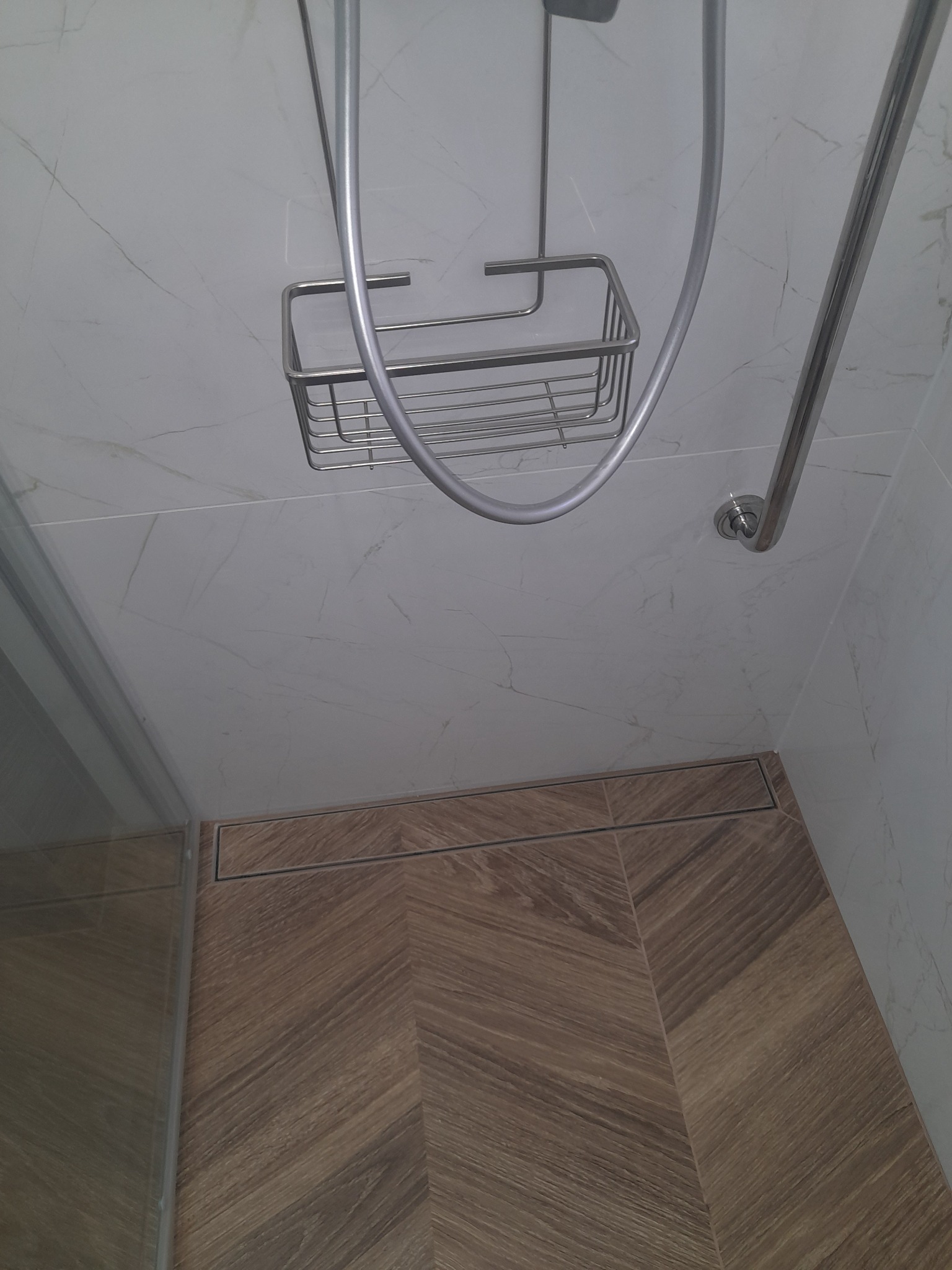 Nowoczesna kabina prysznicowa z odpływem liniowym i półką na kosmetyki. Płytki imitujące drewno na podłodze, marmurowe na ścianach. Minimalistyczny design.