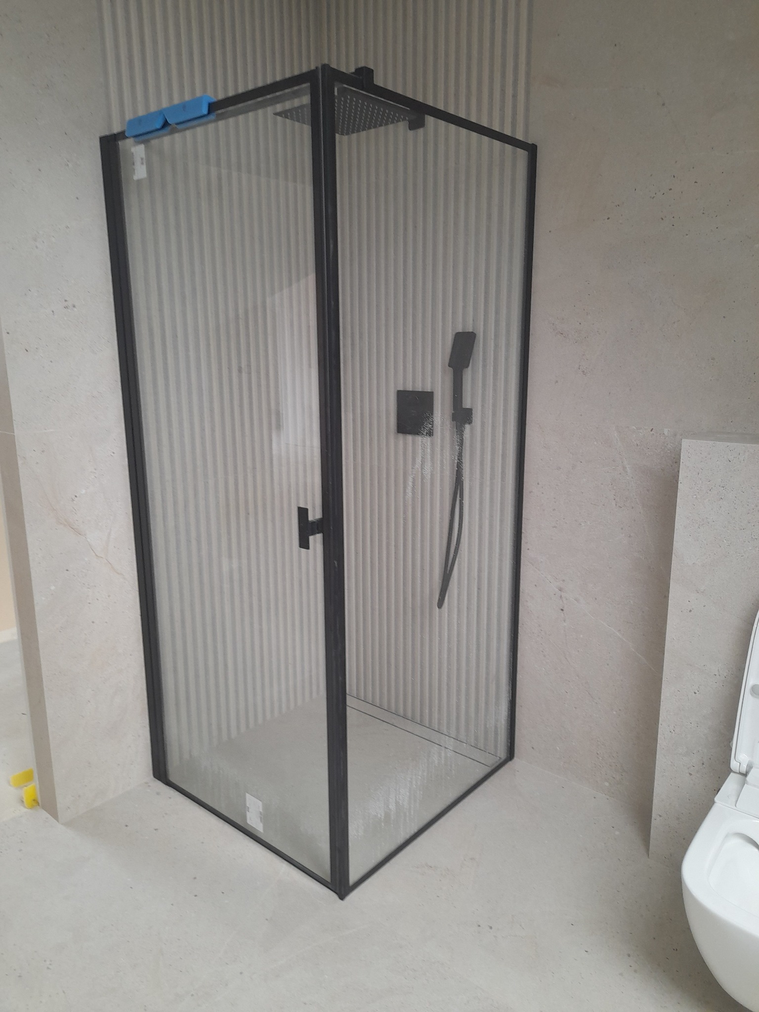 Nowoczesna kabina prysznicowa z czarną ramą, zamontowana w łazience wykończonej płytkami w odcieniach beżu. Widoczne detale: deszczownica i bateria.