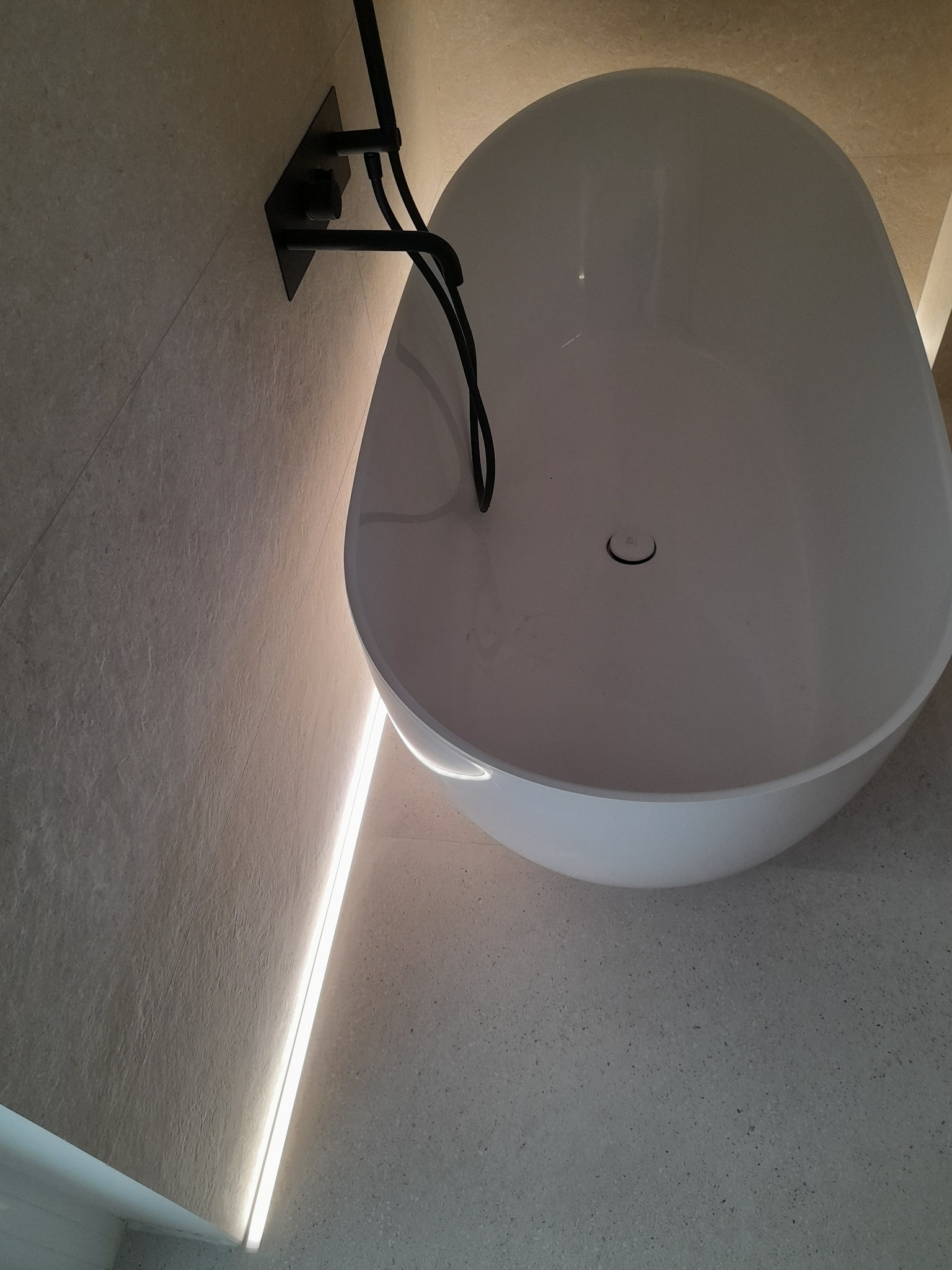 Nowoczesna, wolnostojąca wanna z czarną armaturą na tle ściany wyłożonej płytkami w odcieniach beżu, oświetlona subtelnym światłem LED. Minimalistyczny design.