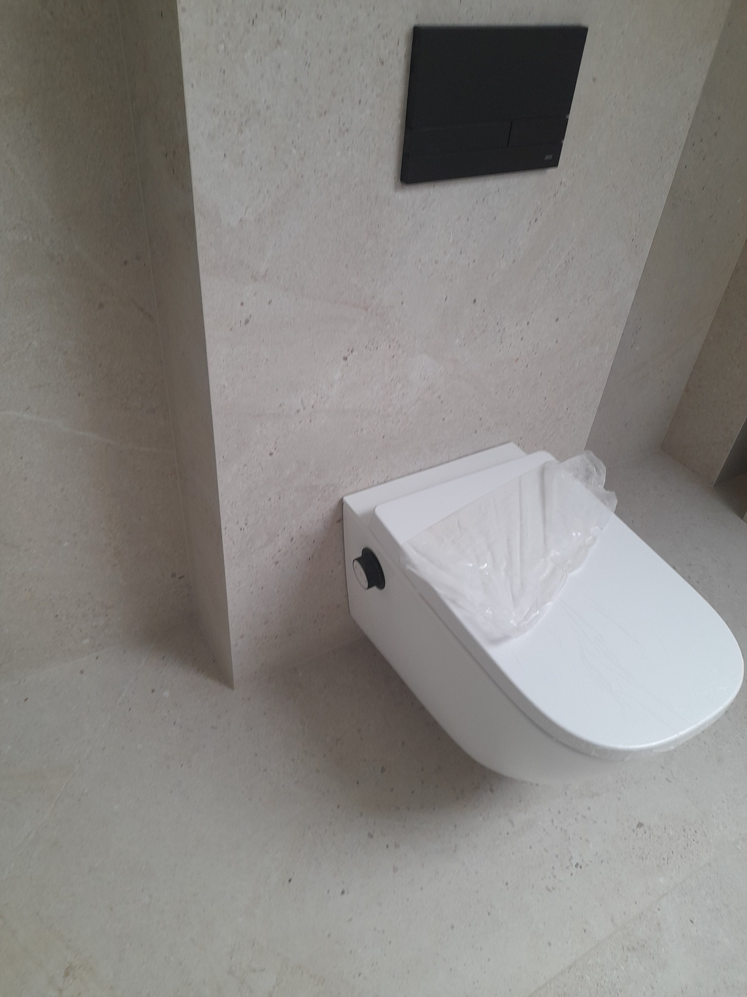 Nowoczesna toaleta podwieszana z czarnym przyciskiem spłukującym na beżowej ścianie z płytek. Toaleta częściowo owinięta folią ochronną.