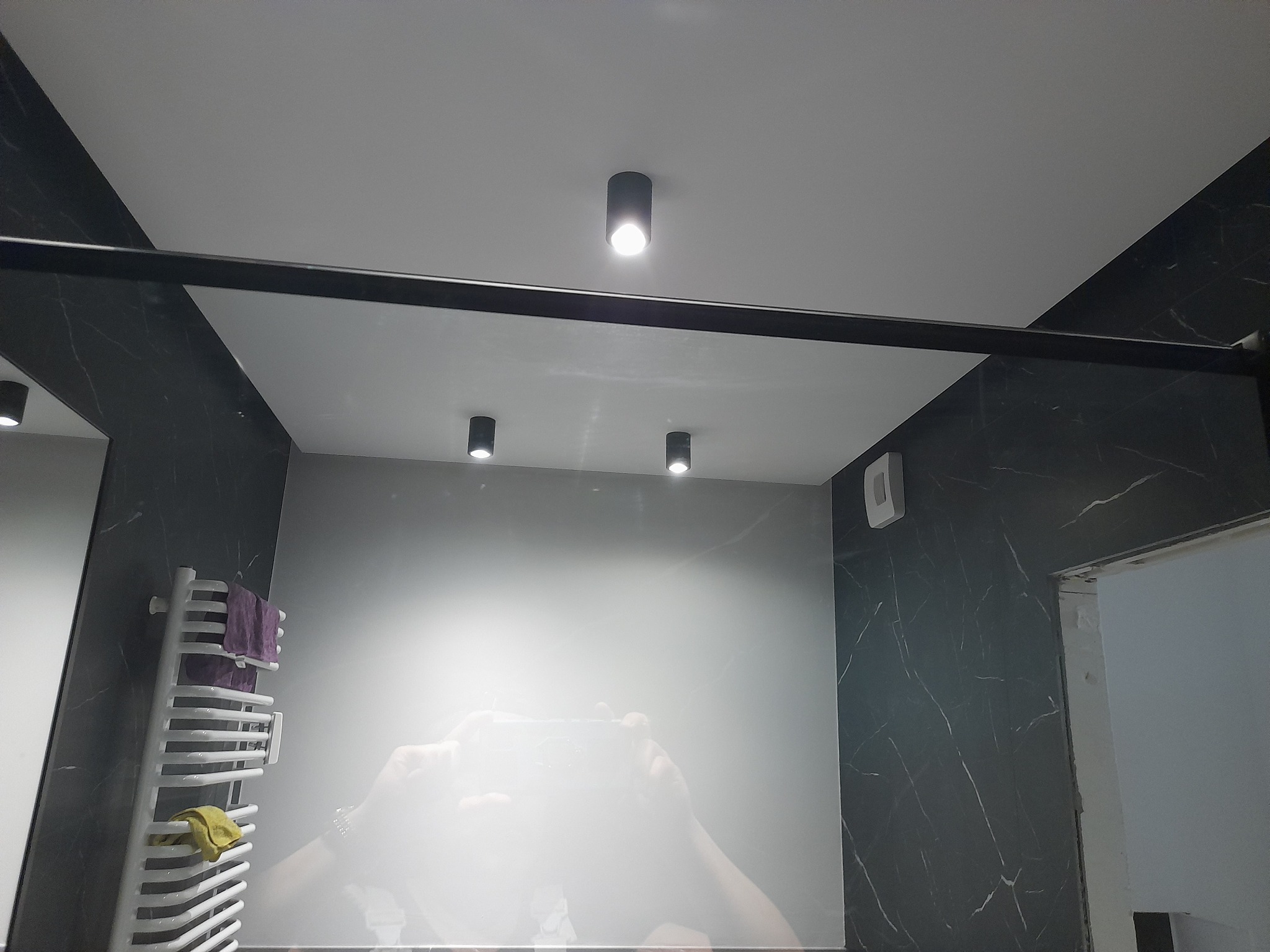 Nowoczesna łazienka z czarnymi marmurowymi płytkami, białym sufitem z trzema punktowymi lampami i grzejnikiem drabinkowym z ręcznikami. Widok z dołu, odbicie fotografa w lustrze.