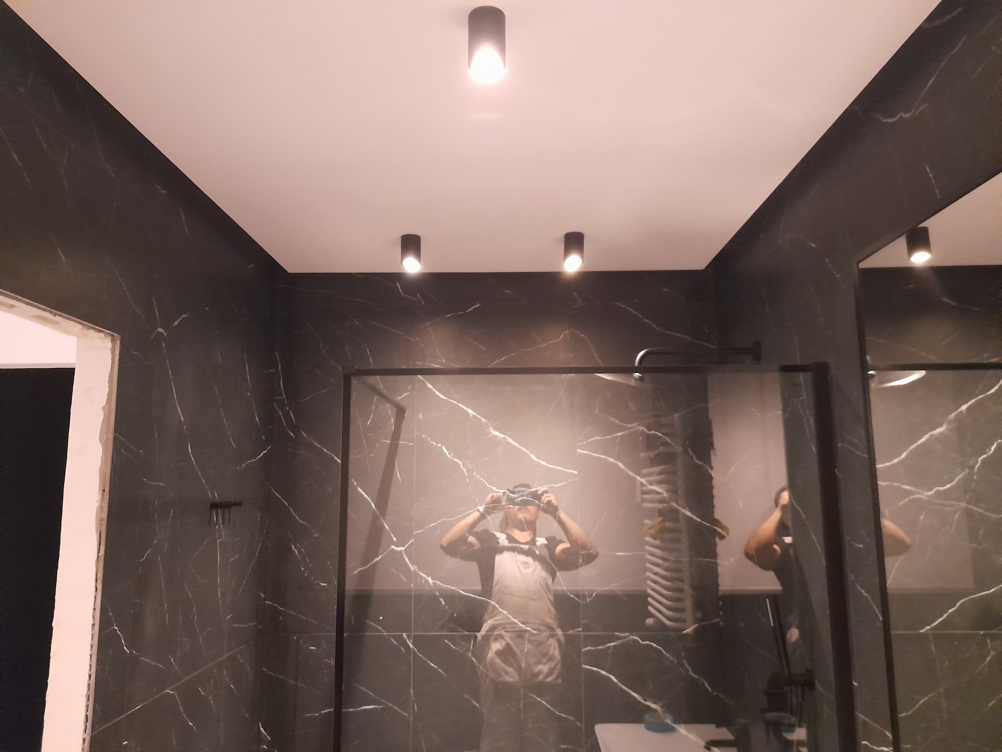 Wykończona łazienka w Zielonej Górze z czarnymi płytkami imitującymi marmur, oświetleniem punktowym i kabiną prysznicową. Widoczne odbicie osoby robiącej zdjęcie.