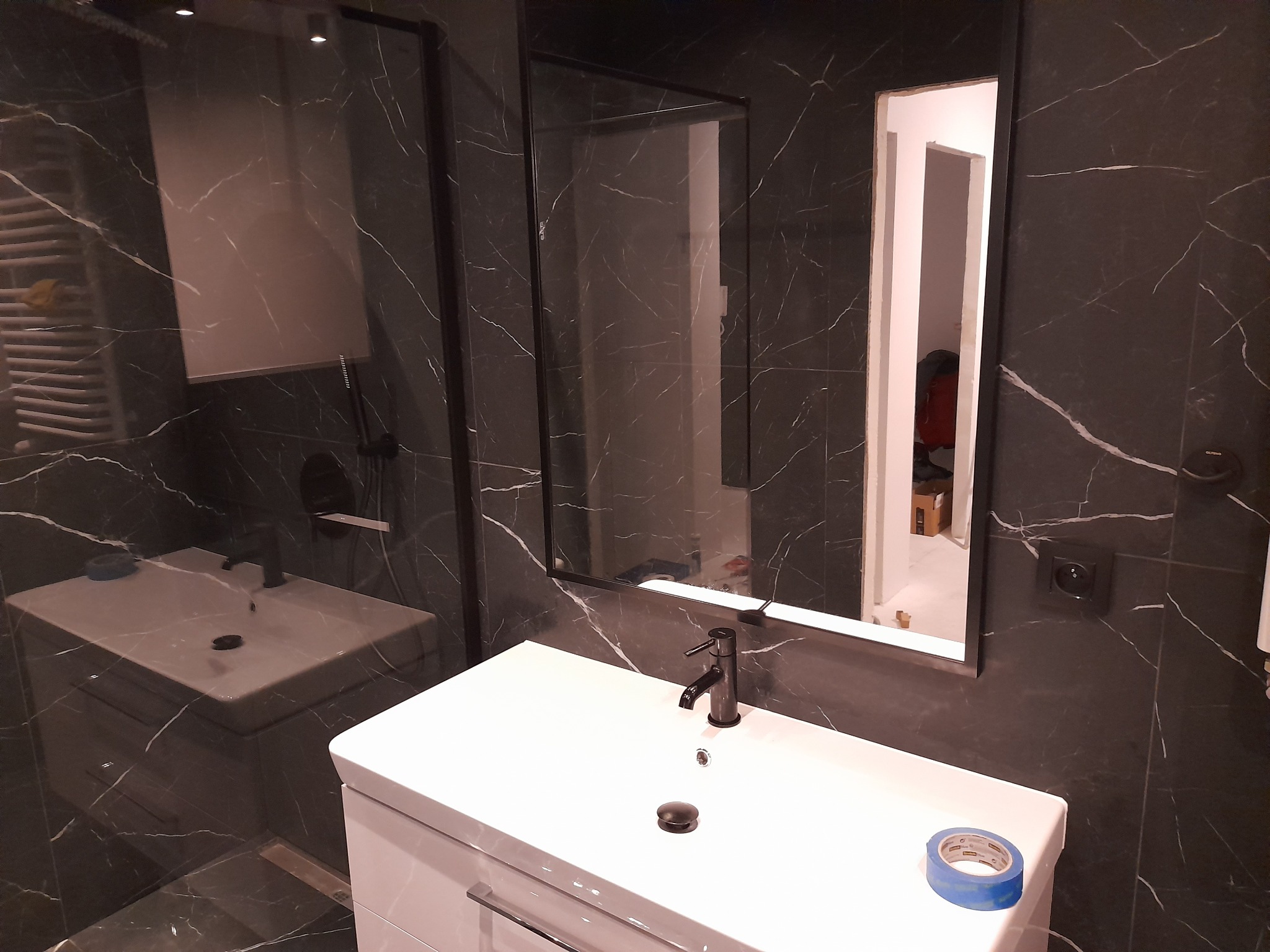 Wykończona łazienka z czarnymi płytkami imitującymi marmur, białą szafką z umywalką i czarną armaturą. W lustrze odbija się fragment kabiny prysznicowej.