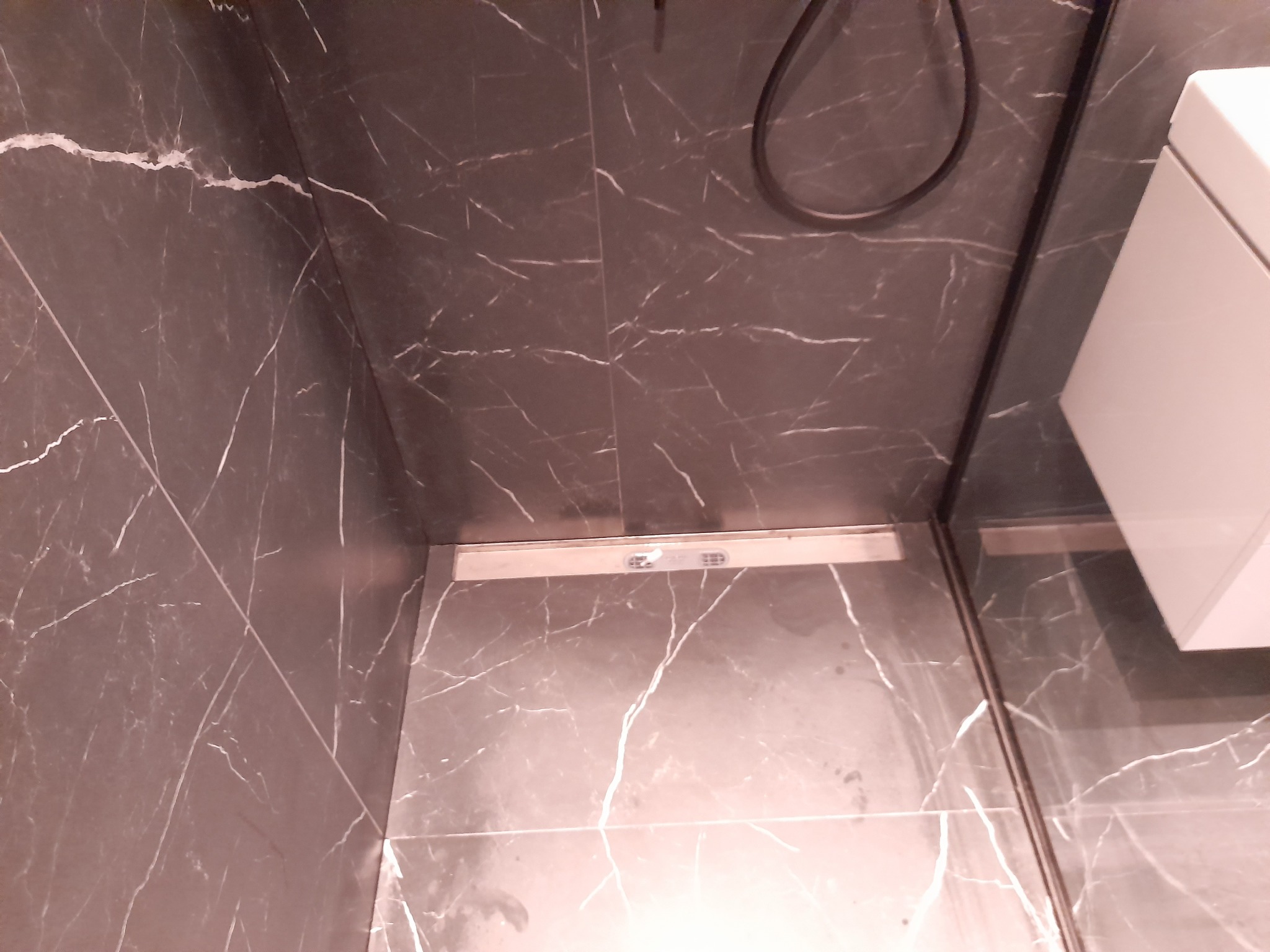 Wykończona łazienka z szarymi płytkami imitującymi marmur, odpływem liniowym i kabiną prysznicową typu walk-in. Widoczny fragment toalety podwieszanej.