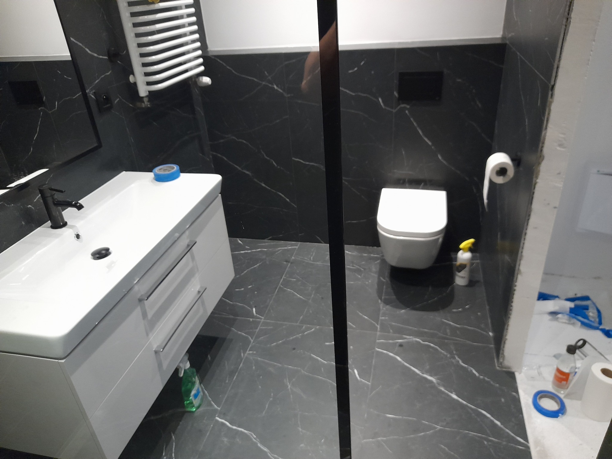 Wykończona łazienka z czarnymi płytkami imitującymi marmur, białą umywalką z szafką i toaletą. Widoczne akcesoria łazienkowe i fragmenty narzędzi glazurniczych.