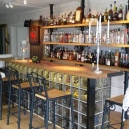 Domowy bar z drewnianym blatem, podświetlanymi luksferami, metalowymi krzesłami barowymi i półkami z alkoholem w tle.