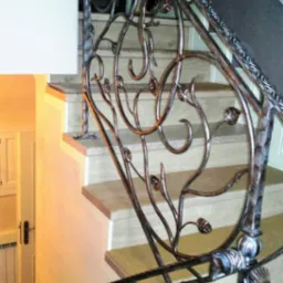 Dekoracyjna balustrada kuta na schodach wewnętrznych z motywem roślinnym, oświetlona ciepłym światłem, z widoczną fakturą metalu i detalami wykończenia.