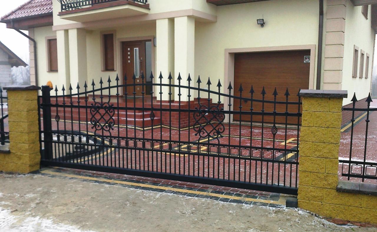 Czarna, metalowa brama przesuwna z dekoracyjnymi elementami, na tle domu z garażem. Żółte słupki z kamienia. Brama zamknięta, widok z zewnątrz.
