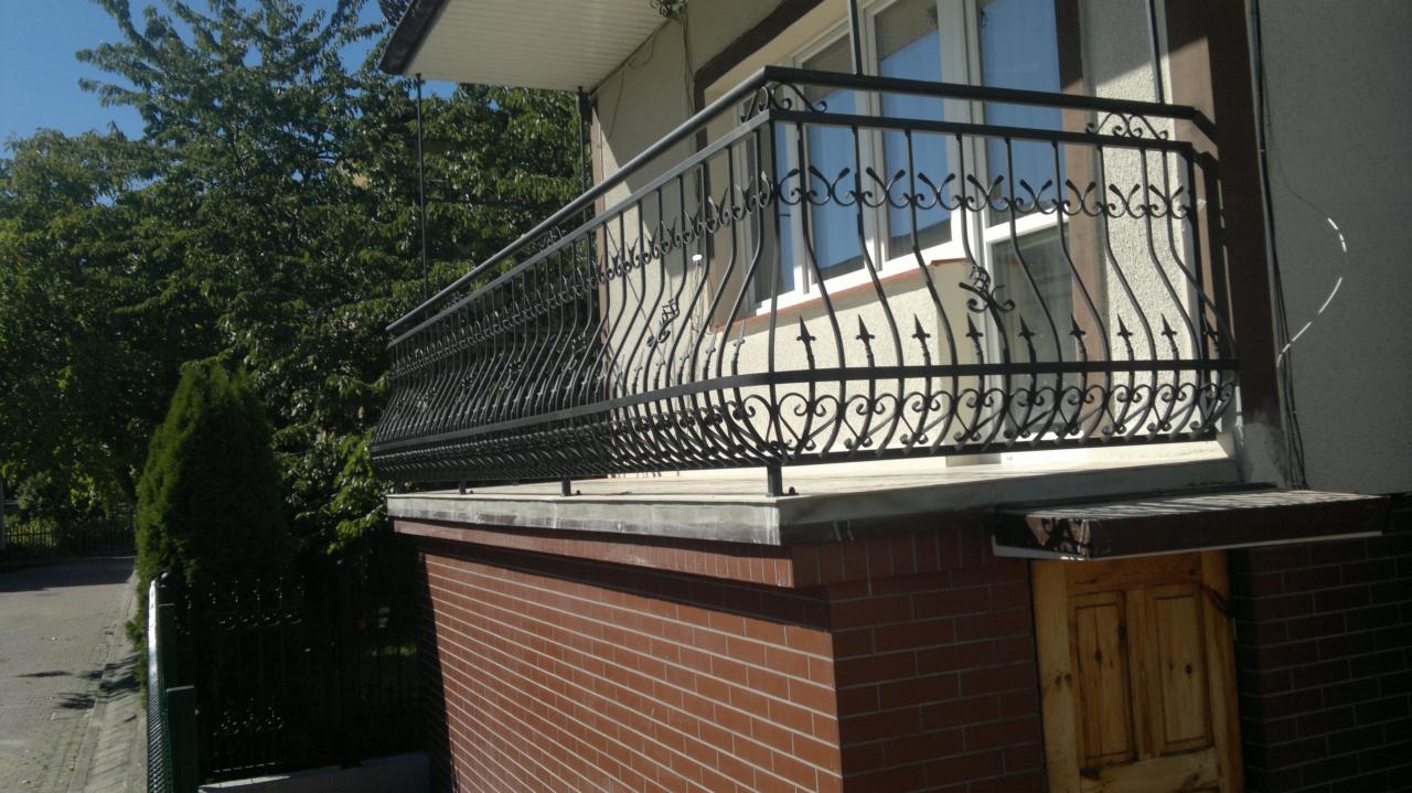 Czarna, metalowa balustrada balkonowa z dekoracyjnymi elementami na tle zieleni i ceglanej ściany budynku. Ujęcie z dołu, podkreślające detale balustrady.