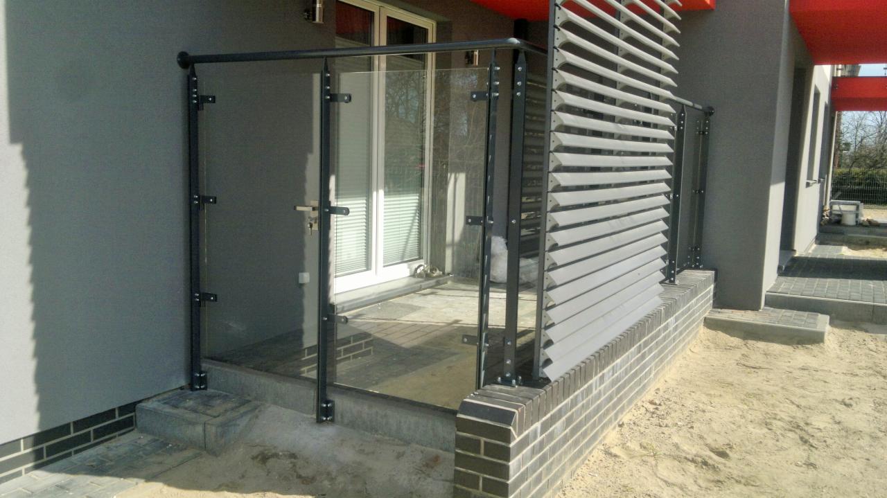 Szklana balustrada balkonowa z czarnymi, metalowymi słupkami, zamontowana przy szarym budynku z szarymi żaluzjami. Widoczny fragment ceglanej podmurówki.